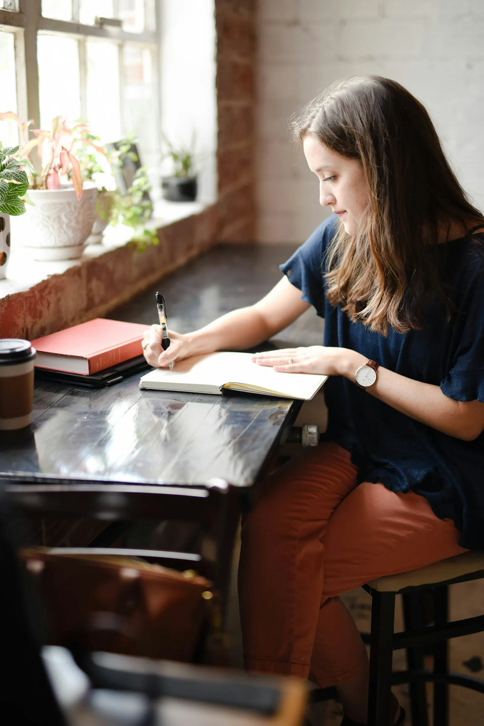 Mulher sentada a uma mesa junto a uma janela, a escrever num caderno, com plantas no parapeito